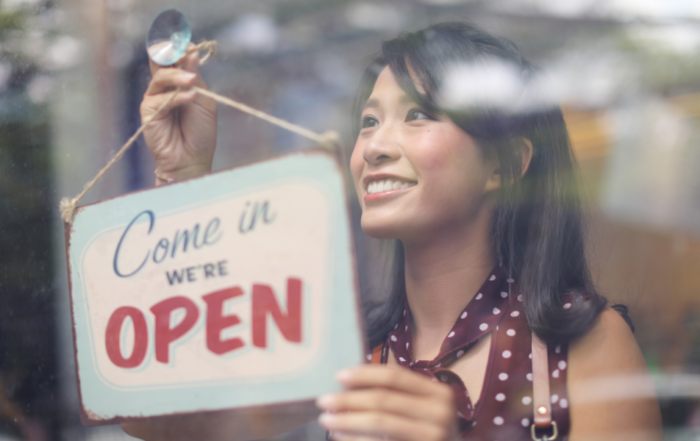 A smiling woman hangs an "Open" sign on a storefront door, inviting customers to come in.