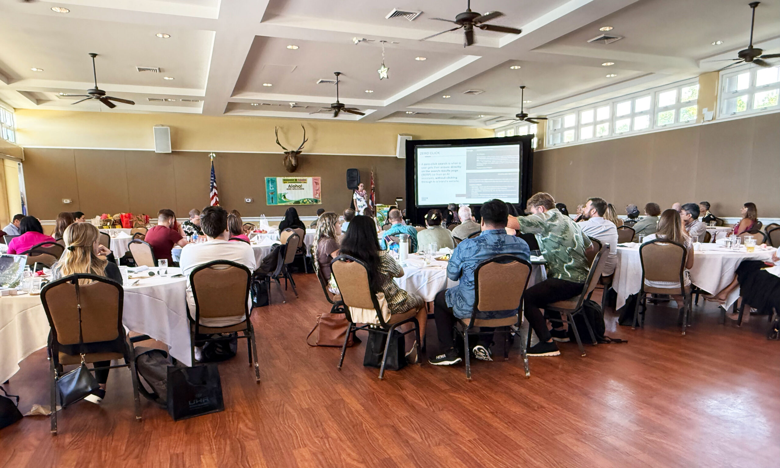AAF-west Anna Covert delivering a keynote presentation for AAF AdWest Hawaii to a seated audience in a conference room, speaking in front of a large screen with attendees gathered at round tables.